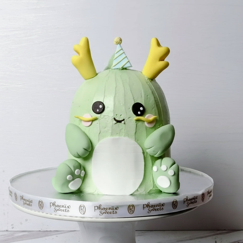 A decorated mini butter cream cake designed to look like a dinosaur, with a green body, white and yellow details, and a 'Happy Birthday' sign on top.