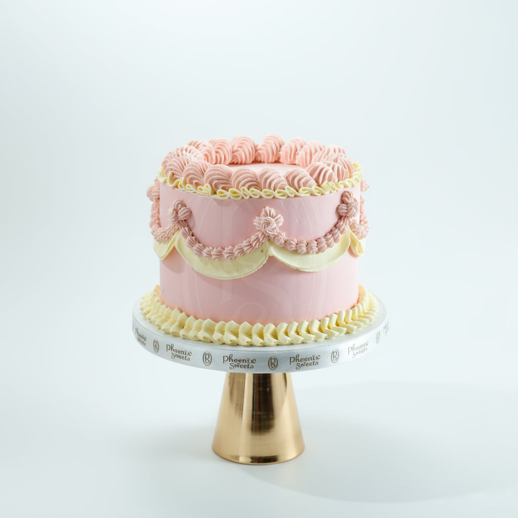 A decorated butter cream cake with pink and yellow tiers, served on a white plate with a gold stand, against a lace background with a floral pattern.