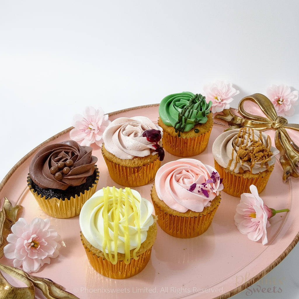 A set of six cupcakes with various toppings and designs, presented on a plate with a floral patterned background. The cupcakes are adorned with decorative elements and there are flowers and a ribbon decoration visible in the background.